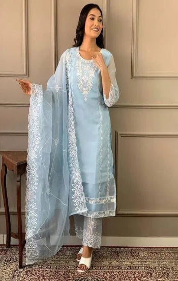 Woman wearing a light blue traditional outfit with white embroidery against a beige wall.